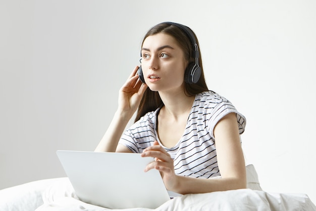 Jolie jeune traductrice travaillant à distance de la maison, assise sur un lit blanc avec ordinateur portable