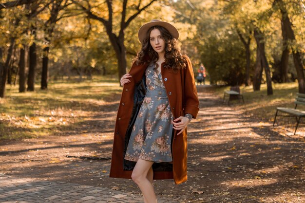 Jolie jolie femme souriante élégante aux cheveux bouclés marchant dans le parc habillé en robe imprimée et manteau chaud automne mode tendance, style de rue
