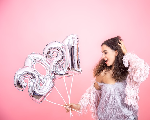 Jolie jolie fille brune aux cheveux bouclés habillée de façon festive posant sur un fond rose avec des ballons d'argent dans les mains pour le concept de nouvel an