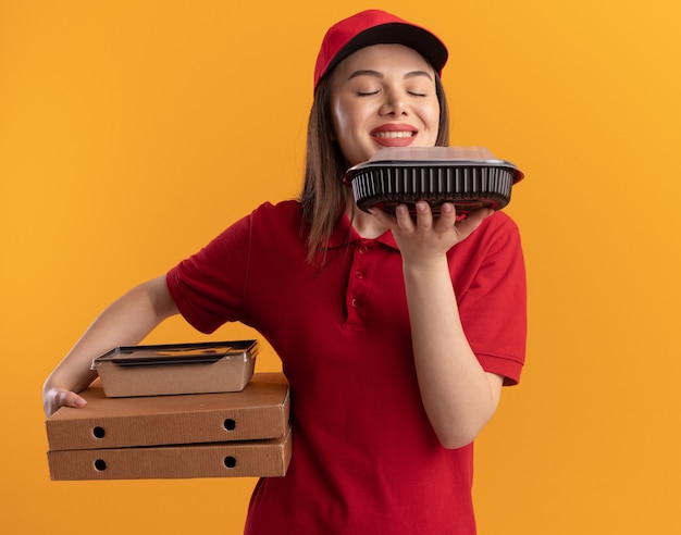 Une jolie livreuse ravie en uniforme tient un emballage alimentaire en papier sur des boîtes à pizza et renifle un contenant de nourriture