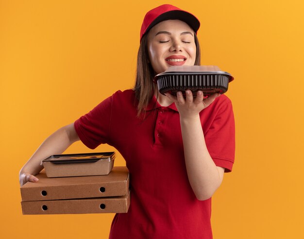 Une jolie livreuse ravie en uniforme tient un emballage alimentaire en papier sur des boîtes à pizza et renifle un contenant de nourriture