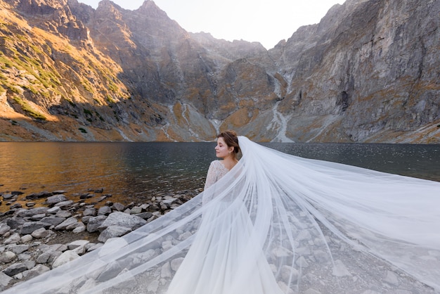 Jolie Mariée Aux Yeux Fermés Et Voile Ondulé Se Tient Devant Le Lac Entouré De Montagnes D'automne Par La Journée Ensoleillée