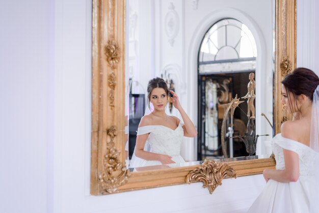 Jolie mariée caucasienne brune regarde dans le miroir de luxe intérieur, jour du mariage