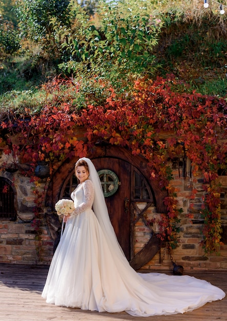 Jolie mariée se tient devant l'entrée en bois d'un immeuble en pierre avec des feuilles colorées de lierre sur la journée ensoleillée