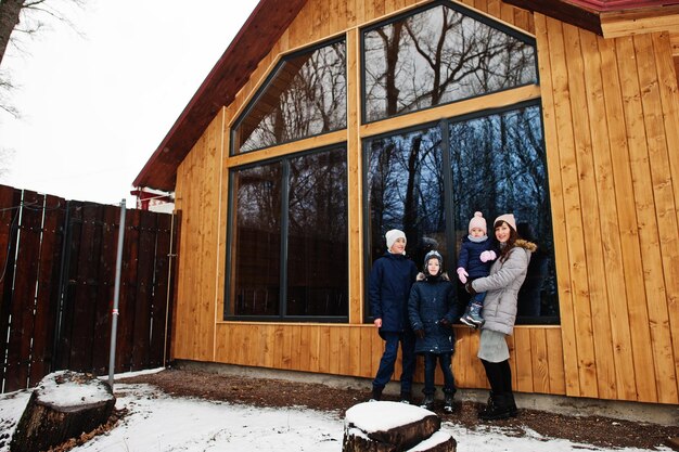 Une jolie mère avec ses enfants en hiver se tient près d'une maison en bois avec une grande fenêtre