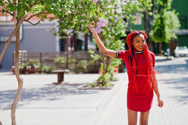 Jolie et mince fille afro-américaine en robe rouge avec des dreadlocks posés en plein air dans la rue près de lilas Modèle noir élégant