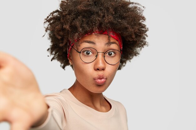 La jolie oman noire fait la moue alors que le selfie étire les mains devant, porte des lunettes transparentes, un pull décontracté