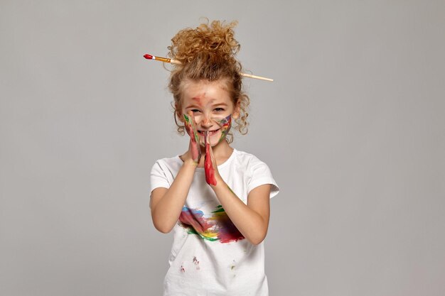 Jolie petite dame ayant un pinceau dans sa jolie coupe de cheveux, vêtue d'un t-shirt blanc enduit. Elle pose avec des mains et des joues peintes, l'air heureuse et souriante, sur un fond gris.