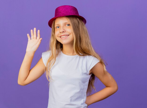 Jolie petite fille au chapeau de vacances en agitant avec la main heureuse et positive, concept de fête d'anniversaire debout sur fond violet