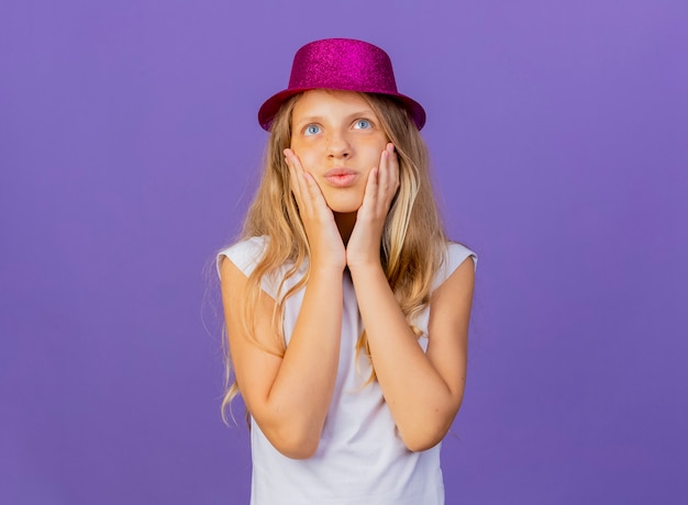 Jolie petite fille au chapeau de vacances touchant ses joues ressentant des émotions positives souriant, concept de fête d'anniversaire debout sur fond violet