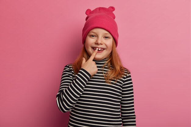 Jolie petite fille au gingembre montre ses deux premières dents adultes, rit et se réjouit, exprime des émotions positives, garde la bouche ouverte, se prépare à un examen oral, vêtue d'un pull rayé et d'un chapeau rose