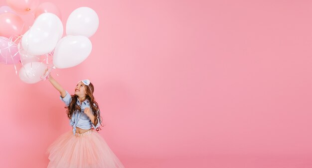 Jolie petite fille aux longs cheveux bouclés, en jupe de tulle rose s'amusant à voler au-dessus des ballons isolés sur fond rose. Bonne enfance d'enfant incroyable exprimant la positivité. Place pour le texte