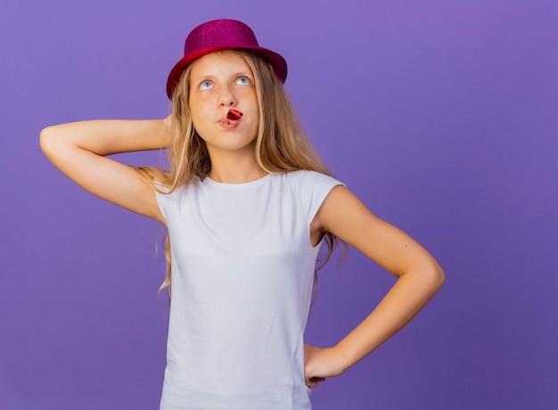 Photo gratuite jolie petite fille en chapeau de vacances blowin whistle à la recherche de concept de fête d'anniversaire perplexe debout sur fond violet