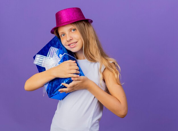 Jolie petite fille en chapeau de vacances étreignant la boîte-cadeau regardant la caméra avec un visage heureux souriant, concept de fête d'anniversaire debout sur fond violet