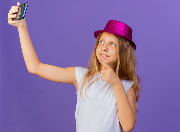 Jolie Petite Fille En Chapeau De Vacances Faisant Selfie à L'aide De Smartphone Souriant Confiant, Concept De Fête D'anniversaire Debout Sur Fond Violet