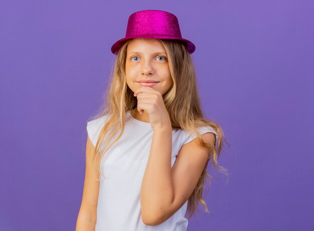 Jolie petite fille en chapeau de vacances regardant la caméra en souriant avec la main sur le menton, concept de fête d'anniversaire debout sur fond violet