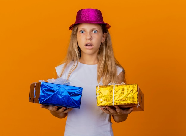 Photo gratuite jolie petite fille en chapeau de vacances tenant des coffrets cadeaux regardant la caméra surpris et étonné, concept de fête d'anniversaire debout sur fond orange
