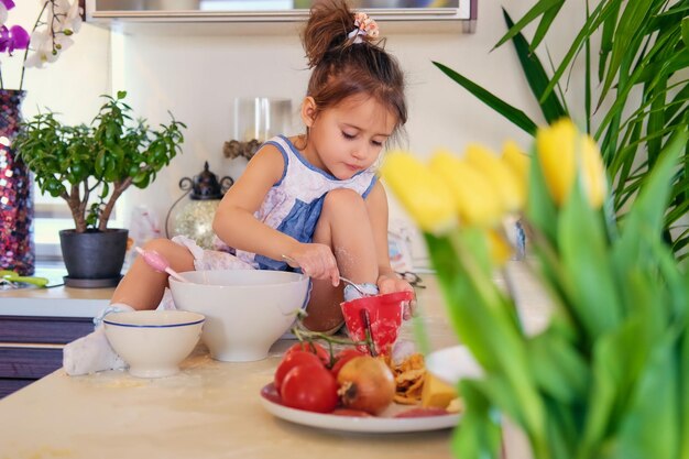 Une jolie petite fille est assise sur une table dans une cuisine et essaie de faire du porridge diététique.