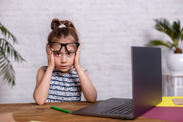 Photo gratuite jolie petite fille est assise à table avec son ordinateur portable et son ordinateur portable, portant des lunettes. le concept de nouveaux programmes pour enseigner aux enfants