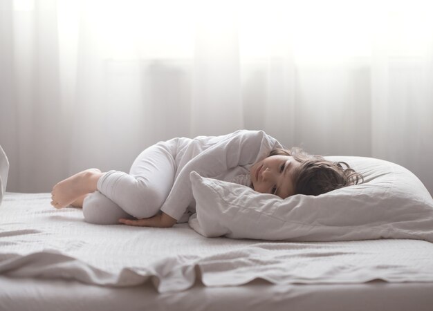 Jolie petite fille est triste couchée dans un lit douillet blanc, le concept de repos et de sommeil des enfants