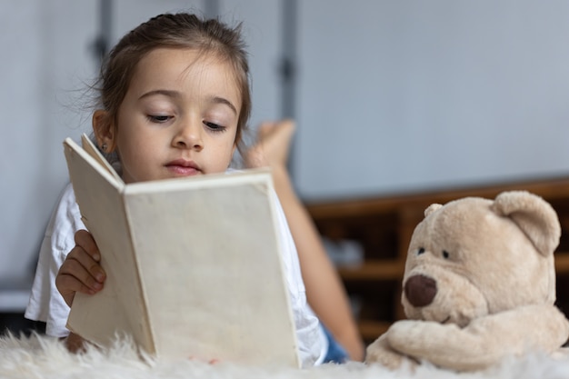 Photo gratuite jolie petite fille à la maison, allongée sur le sol avec son jouet préféré et lit un livre.