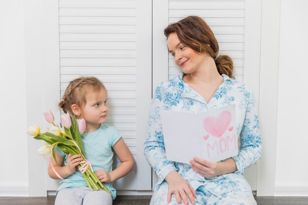 Jolie petite fille et mère se regardant avec une carte de voeux et un bouquet de fleurs de tulipes