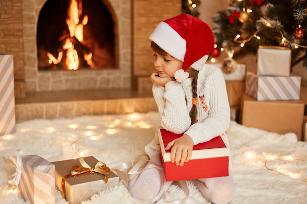 Jolie petite fille pensive portant un pull blanc et un chapeau de père Noël, regardant des boîtes à cadeaux avec une expression faciale pensive, posant dans une salle de fête avec cheminée et arbre de Noël.