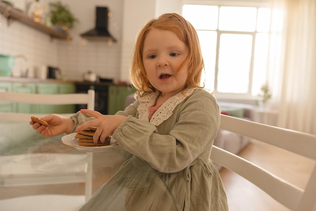 Jolie petite fille rousse caucasienne assise à table en train de manger des biscuits dans des vêtements décontractés pendant la journée à la maison Concept d'aliments pour bébés