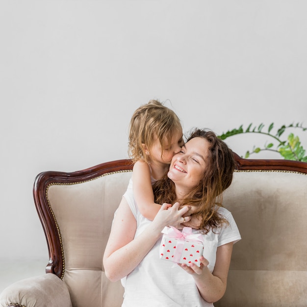 Jolie petite fille s&#39;embrassant à sa mère tenant une boîte-cadeau assise sur un canapé