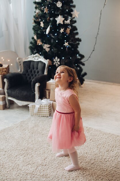 Une jolie petite fille vêtue d'une belle robe s'amuse beaucoup près du sapin de Noël à la maison