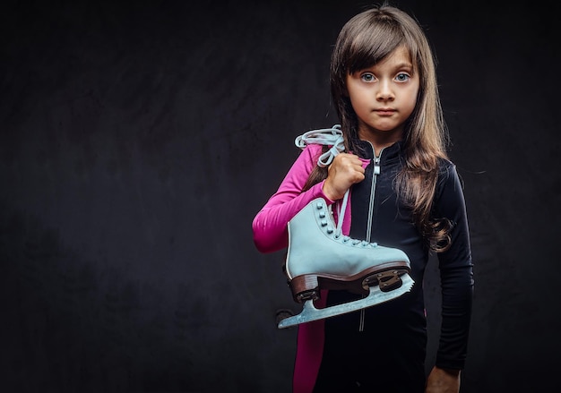 Jolie petite fille vêtue de vêtements de sport tient des patins à glace sur une épaule. Isolé sur un fond texturé sombre.
