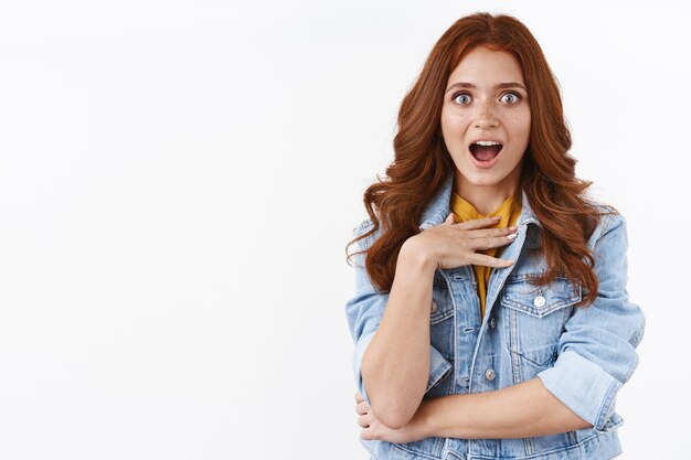 Une jolie rousse surprise en veste en jean, l'air pris dans une embuscade et impressionné, toucher la poitrine avec étonnement, haleter et regarder la caméra étonnée, recevoir un prix inattendu, mur blanc