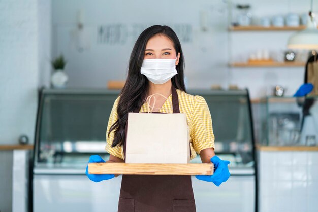 Une jolie travailleuse de café asiatique porte un masque facial et des gants donnant un sac de nourriture à emporter au client