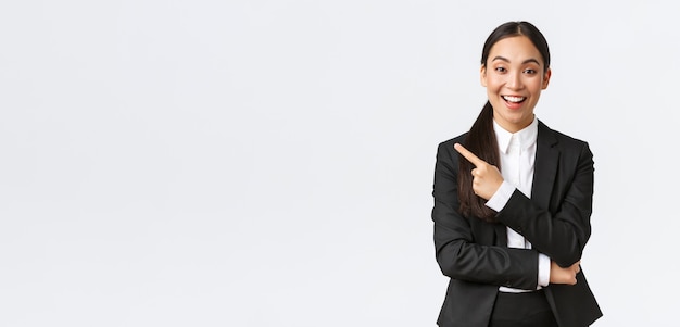 Jolie vendeuse asiatique excitée, agent immobilier en costume suggérant une maison parfaite, debout en costume et pointant le doigt vers la gauche. Femme d'affaires faisant une annonce, afficher la bannière