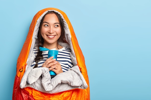 Une jolie voyageuse porte un pull à rayures, enveloppé dans un sac de couchage, tient un thermos avec une boisson chaude, aime le style de vie de camping, a des vacances d'été et de l'aventure, a un charmant sourire à pleines dents sur le visage