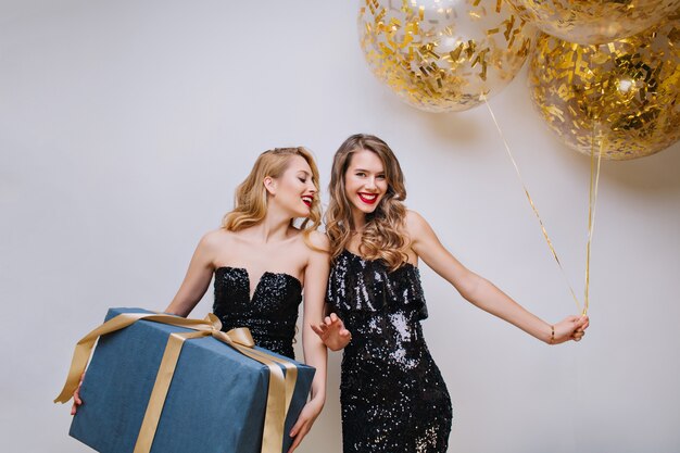 Jolies femmes ypung en robes de luxe noires célébrant la fête d'anniversaire avec un gros cadeau et des ballons. Excité, s'amuser, charmants modèles, célébrer, sourire.