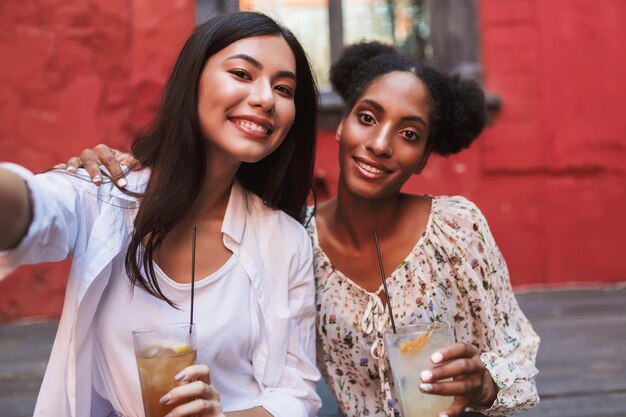 Jolies filles souriantes avec des cocktails dans les mains prenant joyeusement des photos ensemble tout en passant du temps dans la cour du café