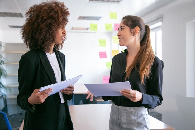 Jolies jeunes femmes d'affaires discutant du plan de projet et souriant. Deux belles collègues féminines tenant des documents et parlant dans la salle de conférence. Concept de travail d'équipe, d'entreprise et de gestion