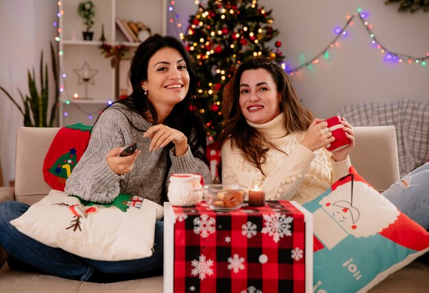 De jolies jeunes filles souriantes tiennent une télécommande de télévision assise sur des fauteuils et profitent de la période de Noël à la maison