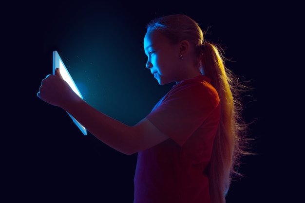 En jouant. Portrait de jeune fille caucasienne isolé sur fond de studio sombre en néon. Beau modèle féminin à l'aide de tablette. Concept d'émotions humaines, expression faciale, ventes, publicité, technologie moderne, gadgets.