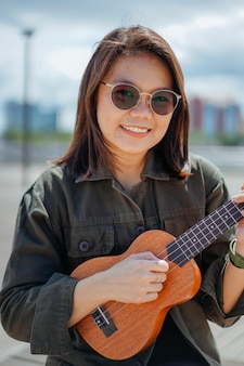 Jouer du ukulélé de la belle jeune femme asiatique portant une veste et un jean noir posant à l'extérieur