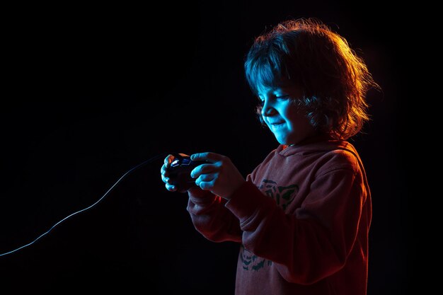 Jouer à des jeux vidéo. Portrait de garçon caucasien sur fond sombre de studio en néon. Beau modèle bouclé.