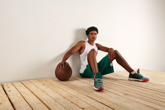 Joueur de basket-ball noir fatigué et réfléchi en basket-ball vert et blanc outift assis sur un plancher en bois clair reposant sa main sur un basket-ball marron grunge