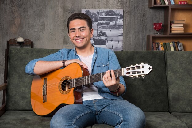 Joueur de guitare souriant tenant une belle guitare et assis sur un canapé. Photo de haute qualité