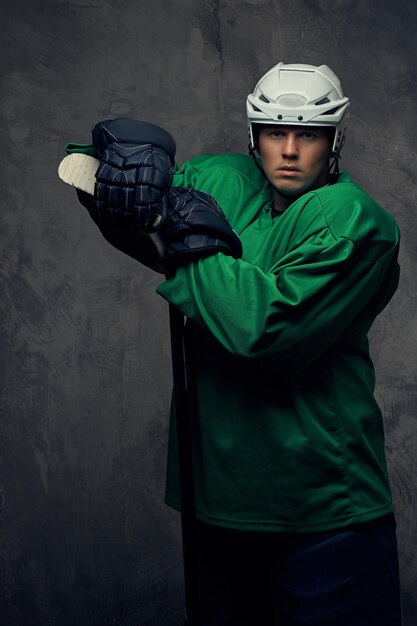 Joueur de hockey portant un équipement de protection vert et un casque blanc debout avec le bâton de hockey sur fond gris.