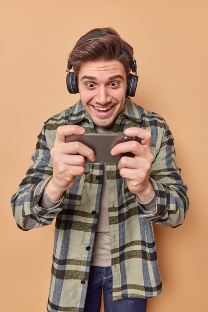 Un joueur optimiste et accro joue à des jeux vidéo sur un smartphone regarde l'écran du cellulaire se sent heureux de porter des écouteurs sans fil sur les oreilles vêtus d'une chemise à carreaux isolée sur fond beige