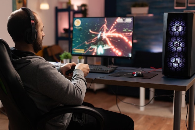Joueur utilisant une manette pour jouer à des jeux vidéo en ligne sur ordinateur. Homme jouant au jeu avec joystick et casque devant le moniteur. Joueur ayant un équipement de jeu, faisant une activité amusante.