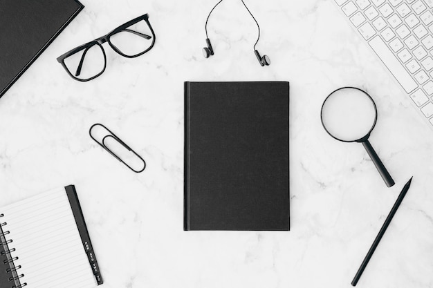 Journal noir fermé avec des lunettes; écouteur; loupes; crayon; clavier et agenda sur le bureau blanc