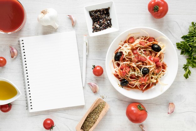 Journal vierge et de délicieuses pâtes spaghettis avec des ingrédients frais sur une table en bois blanche