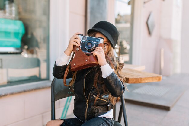 Journée ensoleillée d'heureuse petite fille assise sur la chaise dans le café de la ville et sourire. Elle est élégamment habillée dans ses mains appareil photo rétro. Elle prend des photos de maman, de vraies émotions, de bonne humeur.